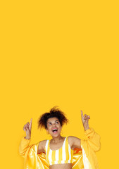 Amazed young african woman wear stylish yellow clothes point fingers look up at copy space, surprised excited black girl show upward advertise fashion sale on summer studio background vertical banner