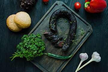 The blood sausage on a dark cutting board is decorated with fresh herbs and vegetables. Blood sausage. View from above. Dark wooden background.