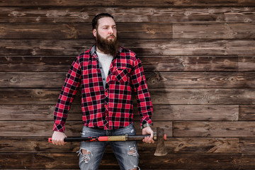 Brutal strong man with a beard dressed in a checked shirt and torn jeans stands with an ax in the hands on the background of a wooden wall