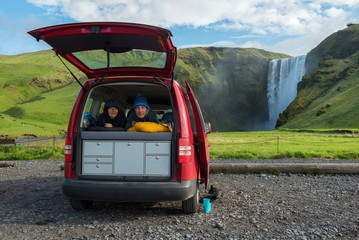 Couple relaxing in a red minivan, Skogafoss waterfall in the background, Iceland