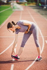 Beautiful young caucasian woman with long hair in tail and big breasts doing exercises, warming up and warming up muscles before training in running stadium, red rubber track in summer on a sunny day