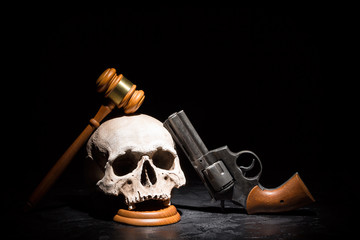 Legal law, justice and murderment crime concept. Wooden judge gavel hammer on human skull with revolver gun pistol against black background