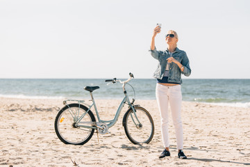 Pretty blonde girl in white pants and denim coat taking selfie on the beach with bicycle. Atrractive woman relaxing near the sea after bike ride