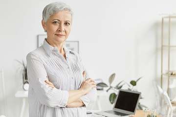 People, age, technolgy and job concept. Good looking serious middle aged female executive with short pixie hairstyle standing at office with arms crossed on chest, her posture expressing confidence