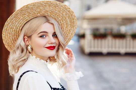 Close up portrait of young beautiful happy lady with red lips makeup, wearing wide brim straw hat, big pearl earrings, posing in street of European city. Copy, empty space for text