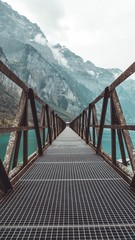 Symmetrische, auf blauen (Berg)see führende Brücke vor einer Bergkette