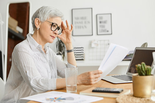 People, Aging, Technology And Profession Concept. Serious 50 Year Old Caucasian Female Wearing Stylish Eyeglasses And Silk Shirt Reading Contract While Working At Desk, Sitting In Front Of Open Laptop