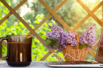 Laptop, cup of coffee, cookies and a basket of lilac flowers on a white wooden table in the summer terrace - concept of remote work, online business, self-employment