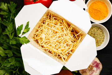 Dry noodles in paper box with greenery, vegetables and spices on a black table, top view