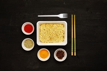 Instant noodles in container with plastic fork, chopsticks and spices on a black wooden table, top view