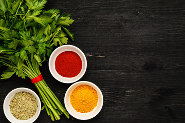 Bunch of green fresh parsley with spices in white ceramic bowls on a black wooden table, top view, text space