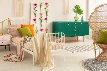 Striped blanket on comfortable single bed in classy bedroom with green wooden cabinet and wicker peacock chair
