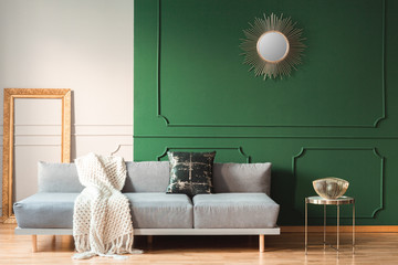 Golden sun shape like mirror on green wall of living room interior with scandinavian sofa with pillows
