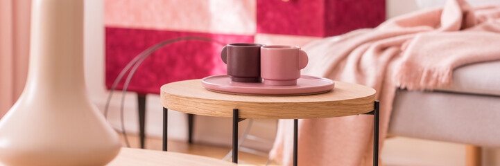 Panoramic view of two pastel pink and burgundy coffee cups on wooden coffee table in elegant pastel pink living room interior
