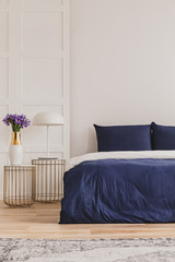 Stylish nightstand with industrial lamp and trendy bedside table with flowers in vase next to simple navy blue bed