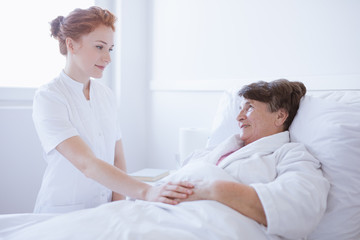 Obraz premium Senior grey woman lying in white hospital bed with young helpful nurse holding her hand