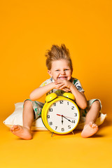 Little cute boy in pajamas holding a toy dinosaur in his hands, sitting on a pillow with an alarm clock. Isolated on a yellow background.