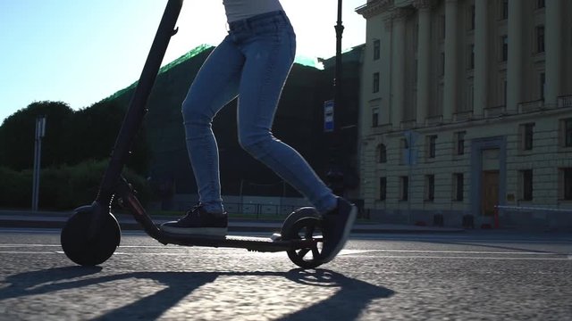 Slow motion - Close up of woman riding electric scooter in a city. Sun lights shine on the vehicle during the moving. 