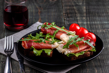 On the table there is a dish with jamon sandwiches, tomato, greens and a glass of red wine