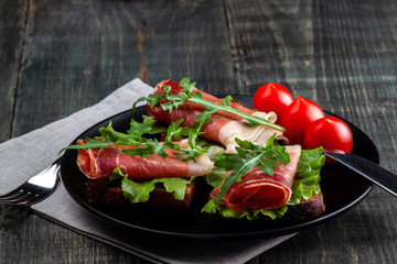 On the table there is a dish with jamon sandwiches, tomato and greens