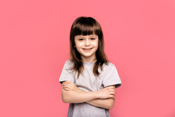 Obraz premium Cheerful little girl keeping arms crossed and looking up with smile while standing against pink background. Confident children concept