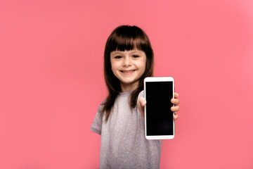 Naklejka premium Surprised happy dark haired little girl kid showing smartphone with blank screen and smiling isolated over pink studio background. 