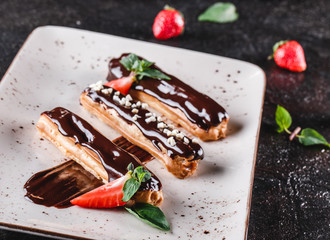 Homemade cake eclairs or profiteroles with custard, chocolate and strawberries on dark background served with cup of coffee. Traditional french eclairs, dessert. Closeup