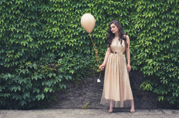 Teen girl in glamorous golden dress standing by the green wall holding balloon. © stivog
