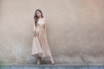 Beautiful teen girl in glamorous golden dress standing by the wall.
