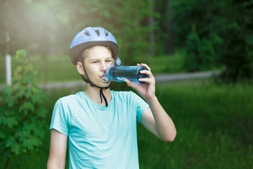 Young boy in helmet and green t shirt cyclist drinks water from bottle in the park. Smiling cute Boy on bicycle in the forest with sport water bottle. Active fun healthy outdoor sport for children. 