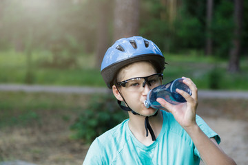 Fototapeta na wymiar Young boy in helmet and green t shirt cyclist drinks water from bottle in the park. Smiling cute Boy on bicycle in the forest with sport water bottle. Active fun healthy outdoor sport for children. 