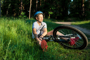Young caucasian boy in helmet and white t shirt got accident and sits on the ground after falling from the bicycle and feels pain on knee in forest in summer. Safety concept, Bicycle accident