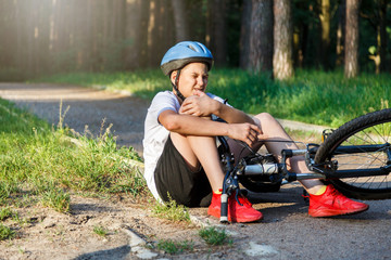 Young caucasian boy in helmet and white t shirt got accident and sits on the ground after falling...