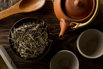 Heap of dried, raw white tea leaves in wooden bowl with teapot and cups on wooden table flat lay top view from above