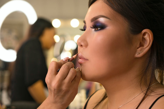 Makeup artist working with Asian model in beauty salon. MUA paints model's lips with contour pencil to make lips more expressive and seductive. Backstage. Photoshoot preparation. Beauty industry