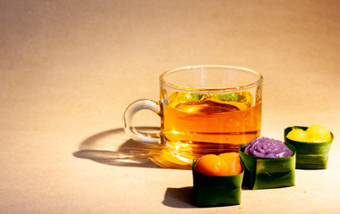 Thai sweets in banana leaves that are arranged with cup of tea on brown paper. Purple rose steamed layer cake, thai mung bean marzipan and golden drops.