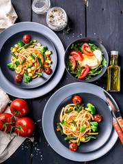 Italian spaghetti with vegetables and salad with arugula and mozzarella on a dark wooden background. Top view.