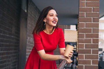 happy beautiful young woman with red lipstick in red dress holding takeaway cup of drink on the street