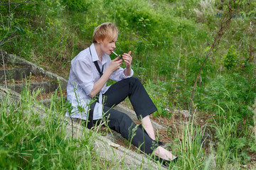 A beautiful and stylish woman with a slightly androgynous appearance with short blond hair (haircut), in a men's white shirt, black pants and tie, sits on the steps and lights a cigarette to smoke