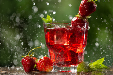in a glass a red drink with strawberries, mint and ice cubes, in backlight, water drops and splashes, summer concept