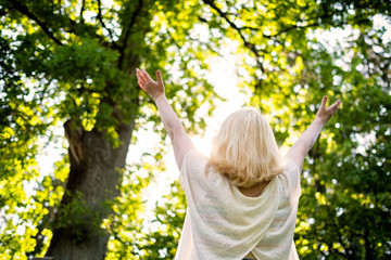 Senior scandinavian woman by the tree holding hands up. Back view