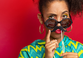 a dark-skinned girl jokes with a cardboard mustache in her face. portrait of funny model on red...