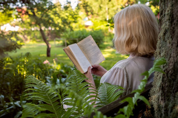 Senior woman in summer garden reading a book sitting near a tree resting. Back side view.