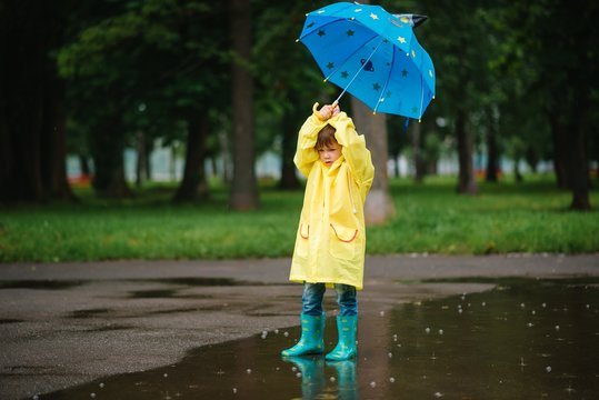 Little boy playing in rainy summer park. Child with umbrella, waterproof coat and boots jumping in puddle and mud in the rain. Kid walking in summer rain Outdoor fun by any weather. happy childhood