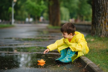 Child playing with toy boat in puddle. Kid play outdoor by rain. Fall rainy weather outdoors activity for young children. Kid jumping in muddy puddles. Waterproof jacket and boots for baby. childhood