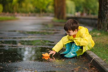 Child playing with toy boat in puddle. Kid play outdoor by rain. Fall rainy weather outdoors activity for young children. Kid jumping in muddy puddles. Waterproof jacket and boots for baby. childhood