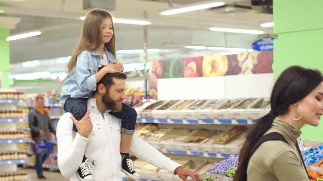 Young fashionable parents buy sweets at the store, the daughter sits on the shoulders of her father and shows her thumb up. Portrait of a happy family in a supermarket. Slow motion.