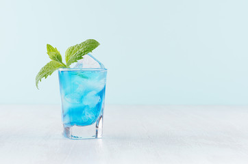 Blue curacao cocktail with ice cubes, green mint in luxury shot glass on white wood board and pastel mint color wall.