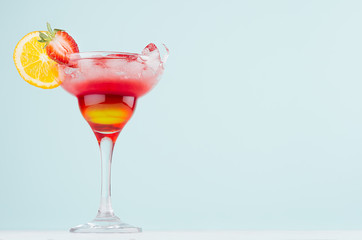 Fresh alcohol striped cocktail with red and yellow liquor, orange slice, strawberry, ice on elegant pastel mint color background and white wood board.