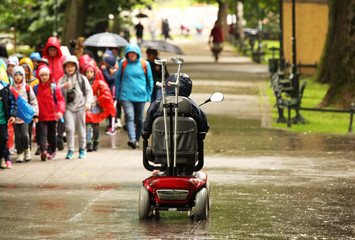 An elderly man in a mechanical wheelchair is passing by the alee of the park past a passing group of children. Sympathy and help to people in need. Active and socialization of persons with disabilitie